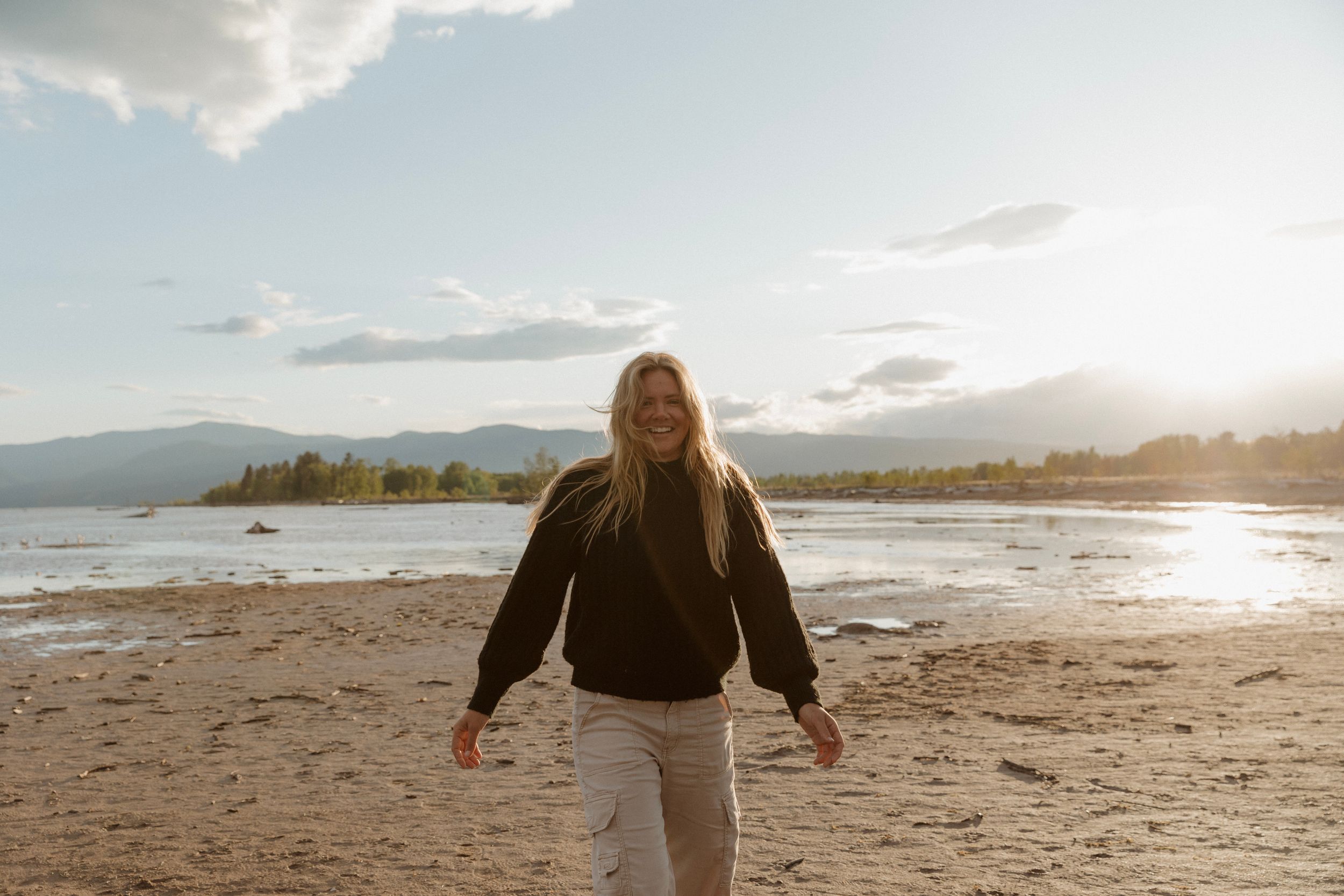 A smiling woman with long blonde hair walks on a sandy beach at golden hour with mountains in the background.