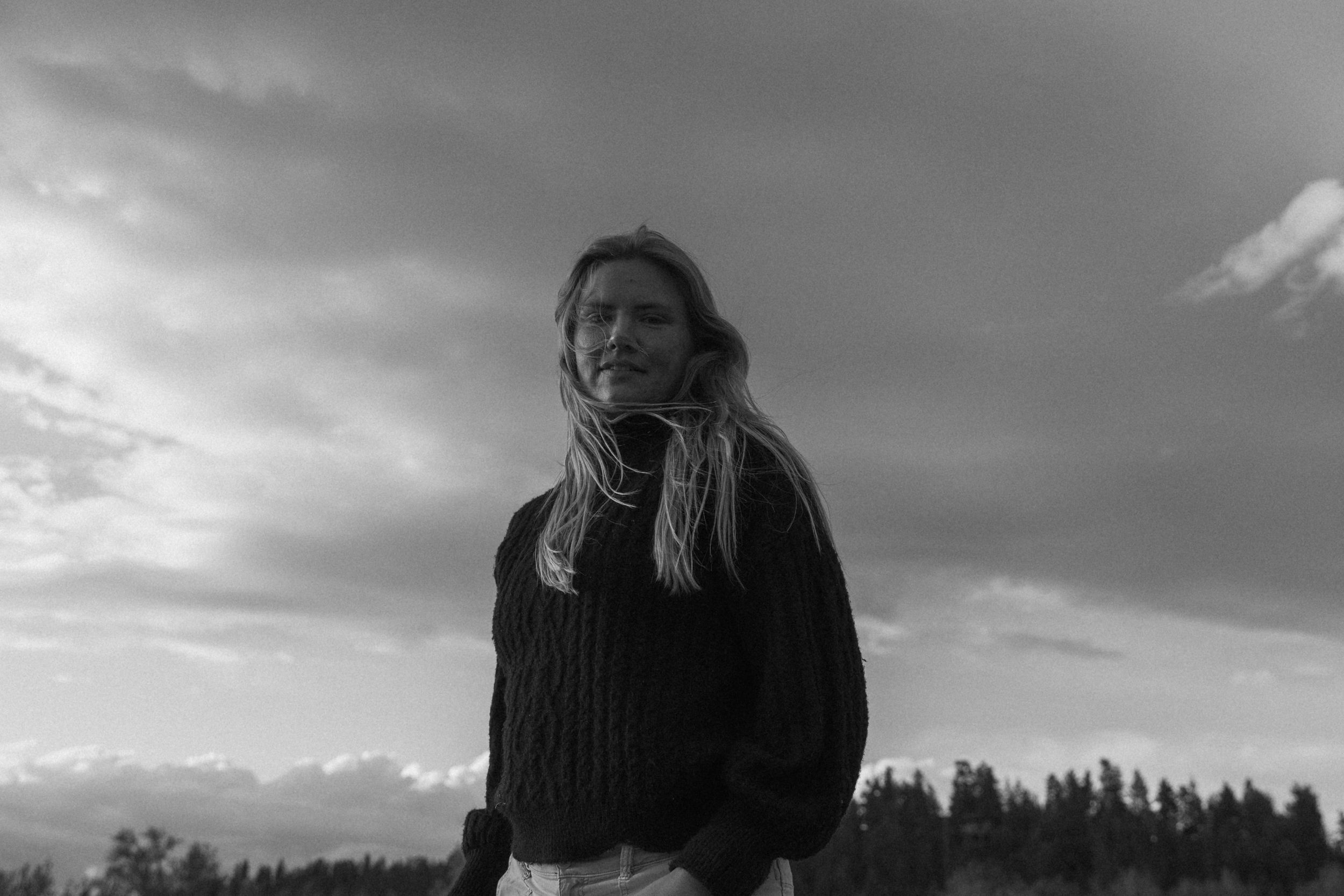 Black and white photo of a young woman in a knit sweater standing outdoors under a dramatic cloudy sky.
