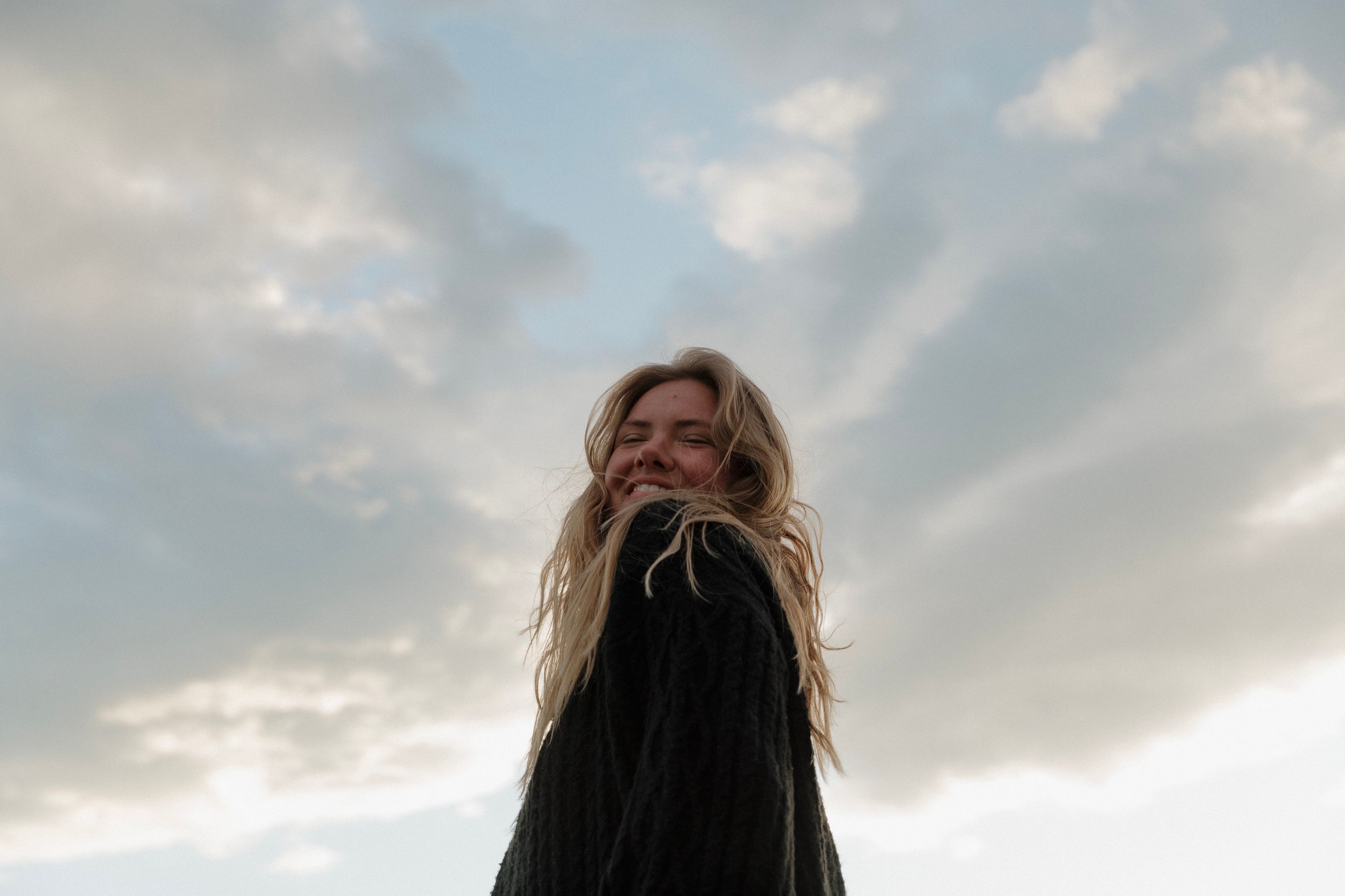 Smiling blonde woman with windswept hair against a dramatic cloudy sky, shot from below in a moody outdoor portrait.