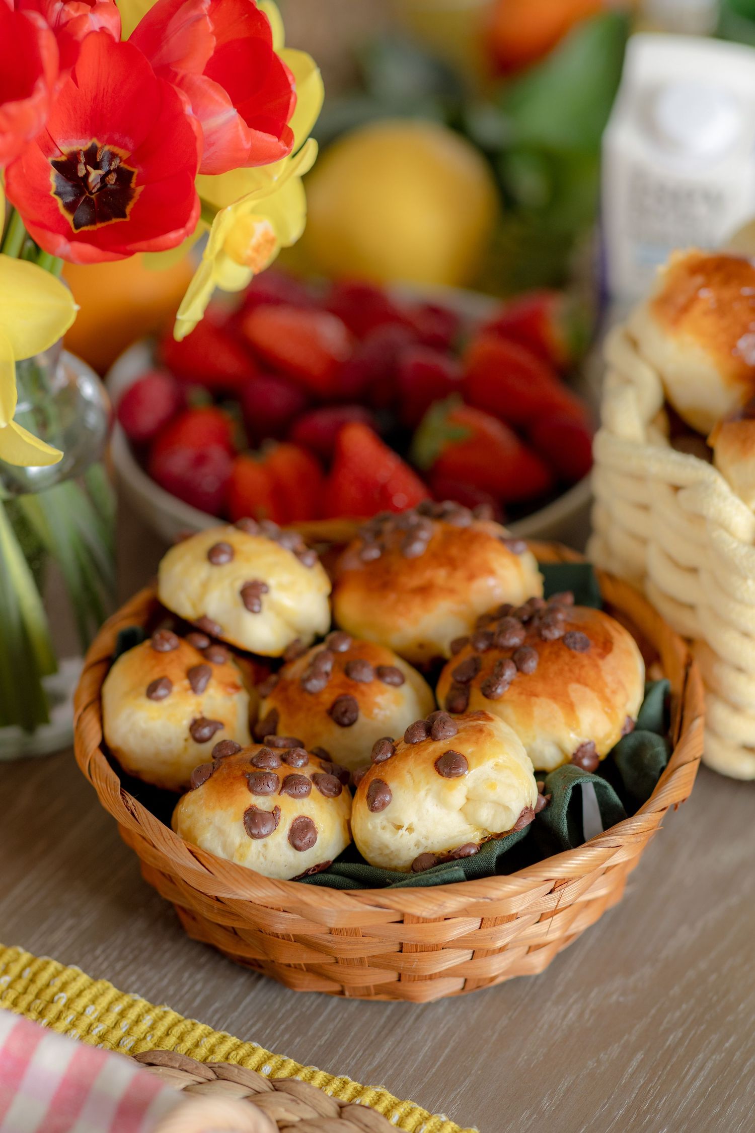 Wicker basket filled with chocolate chip rolls beside fresh strawberries and tulips on a wooden table.