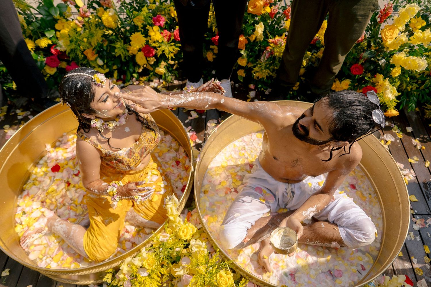 Couple in golden tubs filled with flowers and petals during a vibrant Indian haldi pre-wedding ceremony.