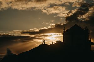 Dramatic sunset silhouettes against cloudy sky with mountains and architectural elements in panoramic view.