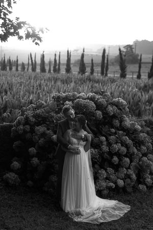 A romantic black and white wedding portrait with a couple embracing by a hydrangea bush with cypress trees.
