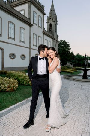 A romantic wedding couple embraces outside a historic church with stone architecture and manicured grounds.