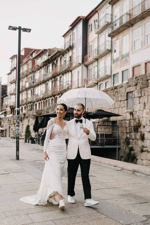 Newlyweds share a laugh while walking with white umbrella along historic European street.