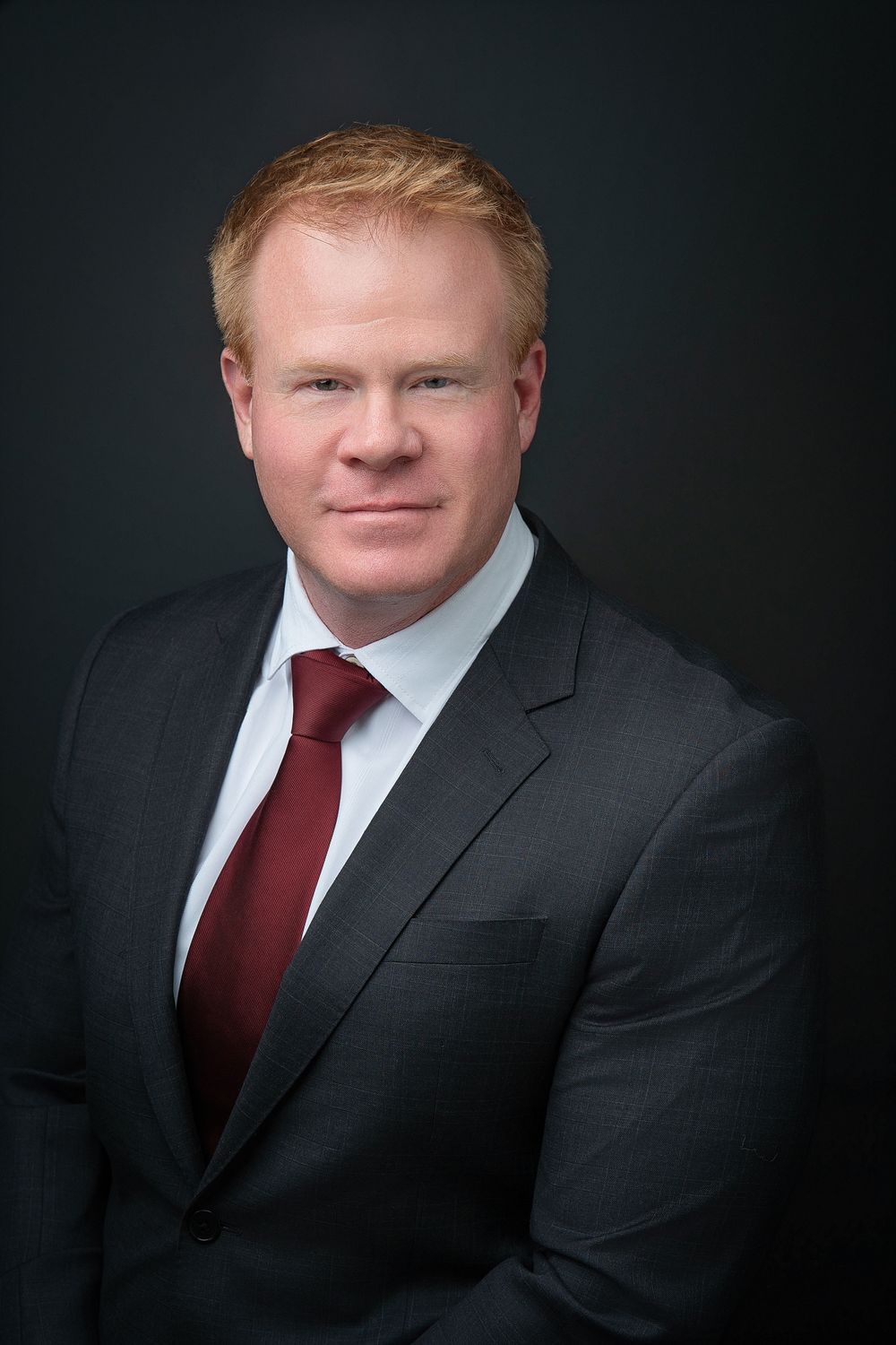 Professional headshot of a red-haired man in a gray suit and burgundy tie against a dark background.