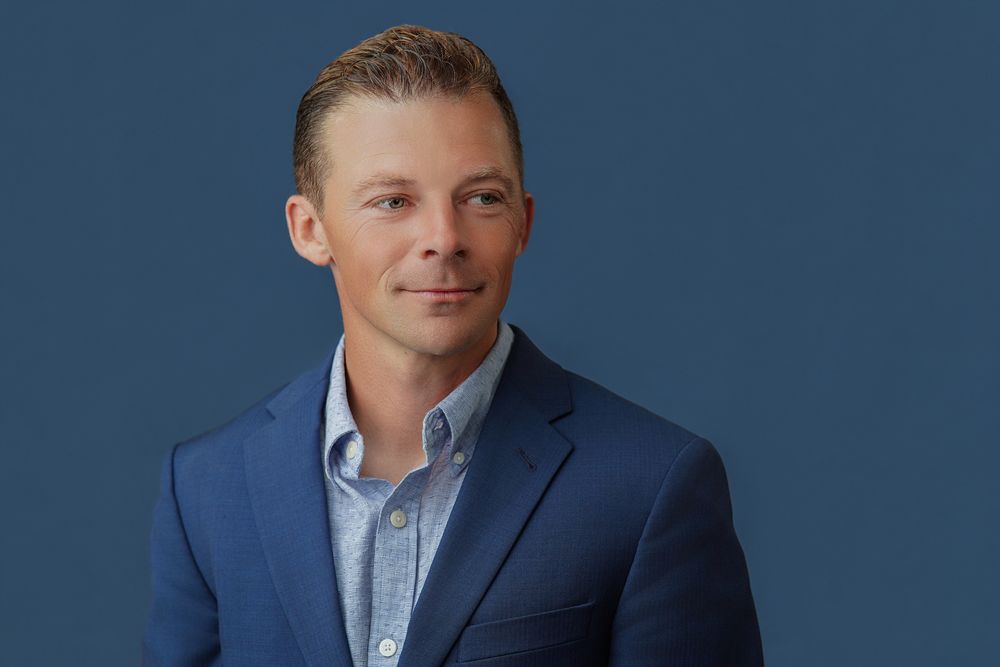 Professional businessman in blue blazer and collared shirt posing against a dark blue background.