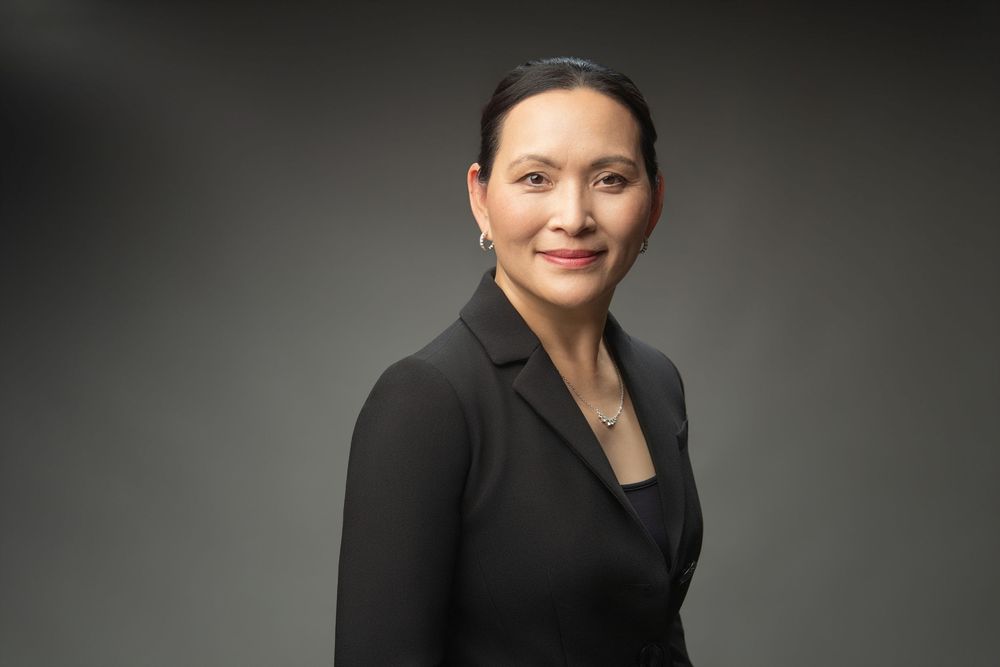Professional woman in black blazer smiling confidently against gray background in corporate headshot photo.
