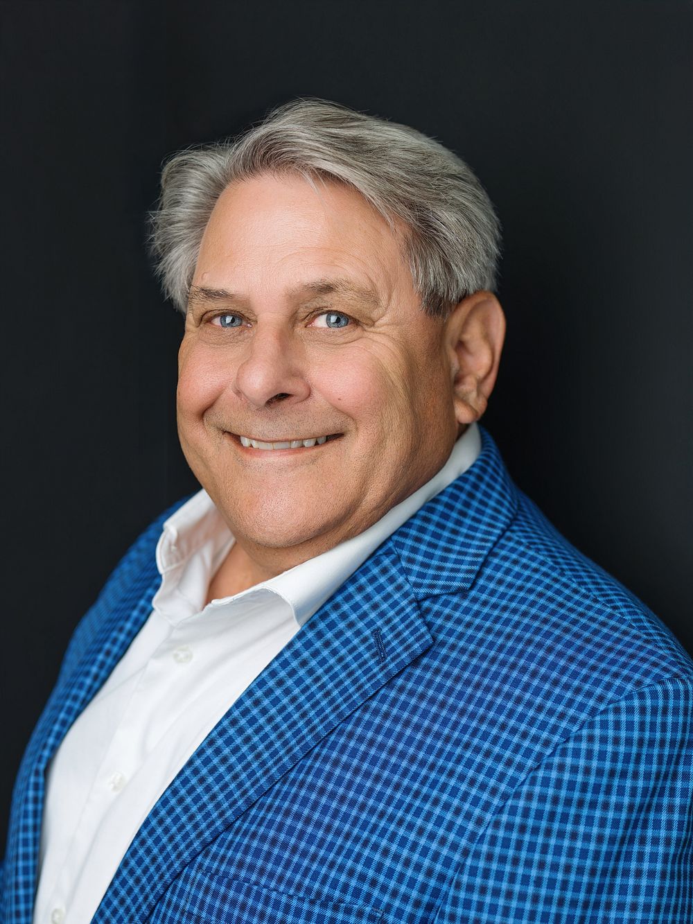 Professional headshot of a smiling middle-aged man with gray hair wearing a blue plaid blazer and white shirt.