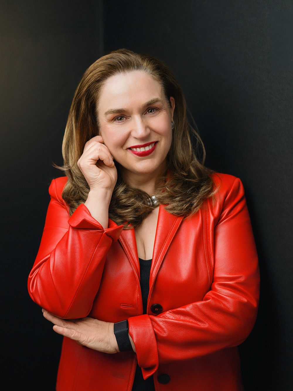 Woman in red leather blazer smiling confidently, posing with hand near chin against dark background.