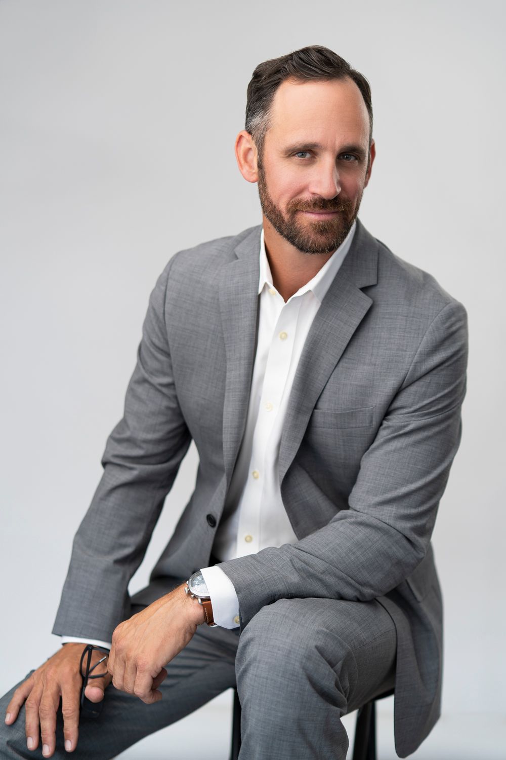 Professional man in gray suit sitting confidently against white background for business headshot photo.