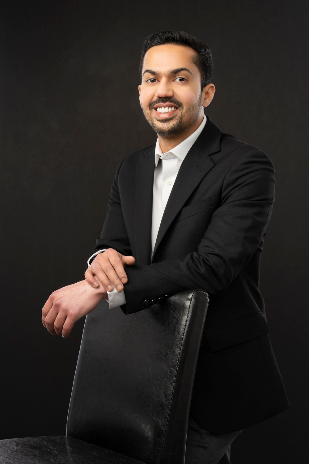 Smiling man in black suit leaning on chair against dark background, professional portrait photo.