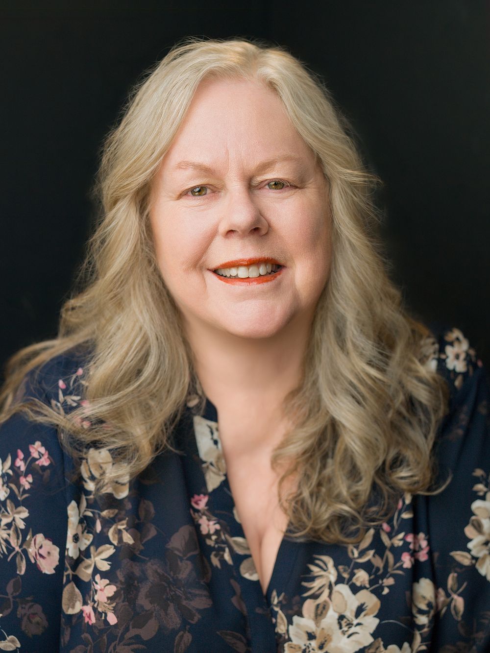 Smiling middle-aged woman with long blonde wavy hair wearing a floral navy blouse against a dark background.