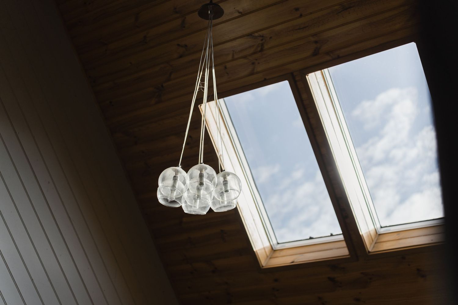 Rustic wooden ceiling with skylight windows and hanging cluster of clear glass globe pendant lights against blue sky.
Cedar & Stone