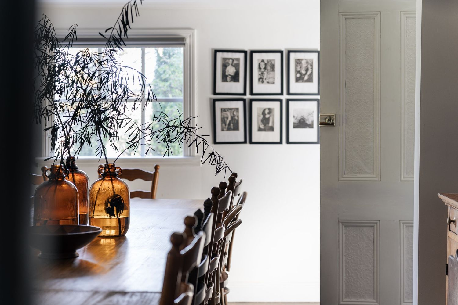 Rustic dining room with long wooden table, ladder-back chairs, black-and-white gallery wall, and olive branch.

Piccadilly House