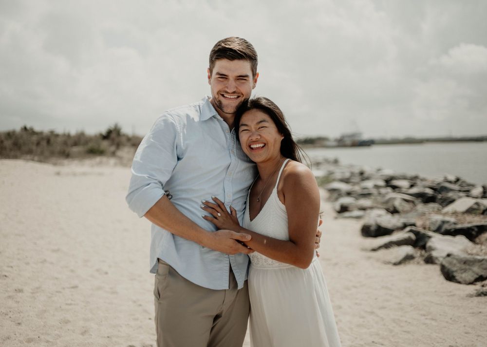 Happy couple embracing on a sandy beach with rocky shoreline, man in blue shirt and woman in white dress.