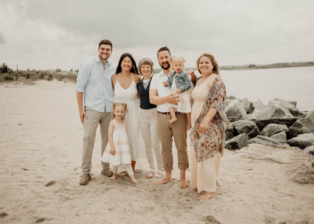 Family of seven posing together on a sandy beach with rocky jetty backdrop on a cloudy day.