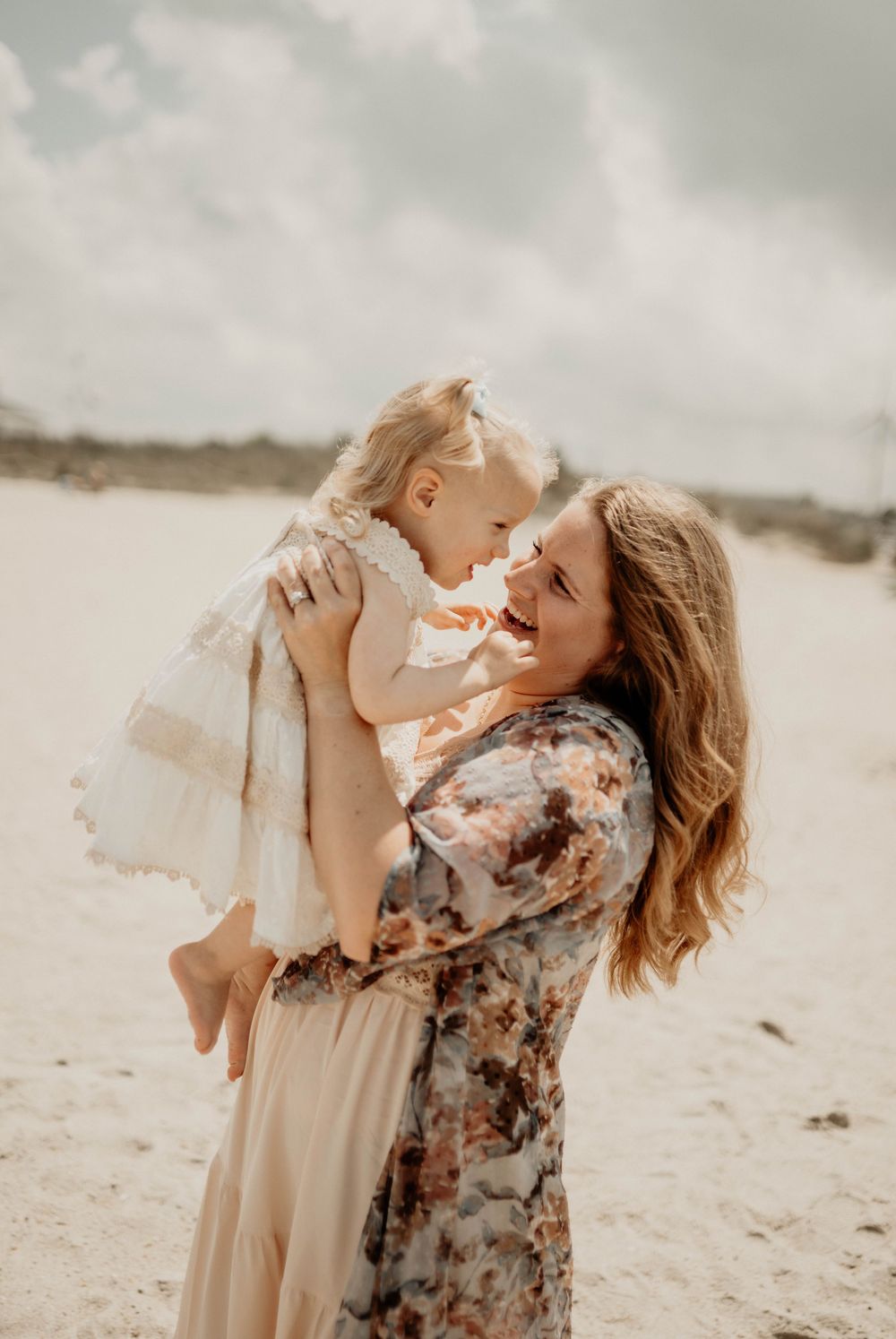 A joyful mother lifts her smiling toddler girl on a sandy beach, both laughing together on a cloudy day.