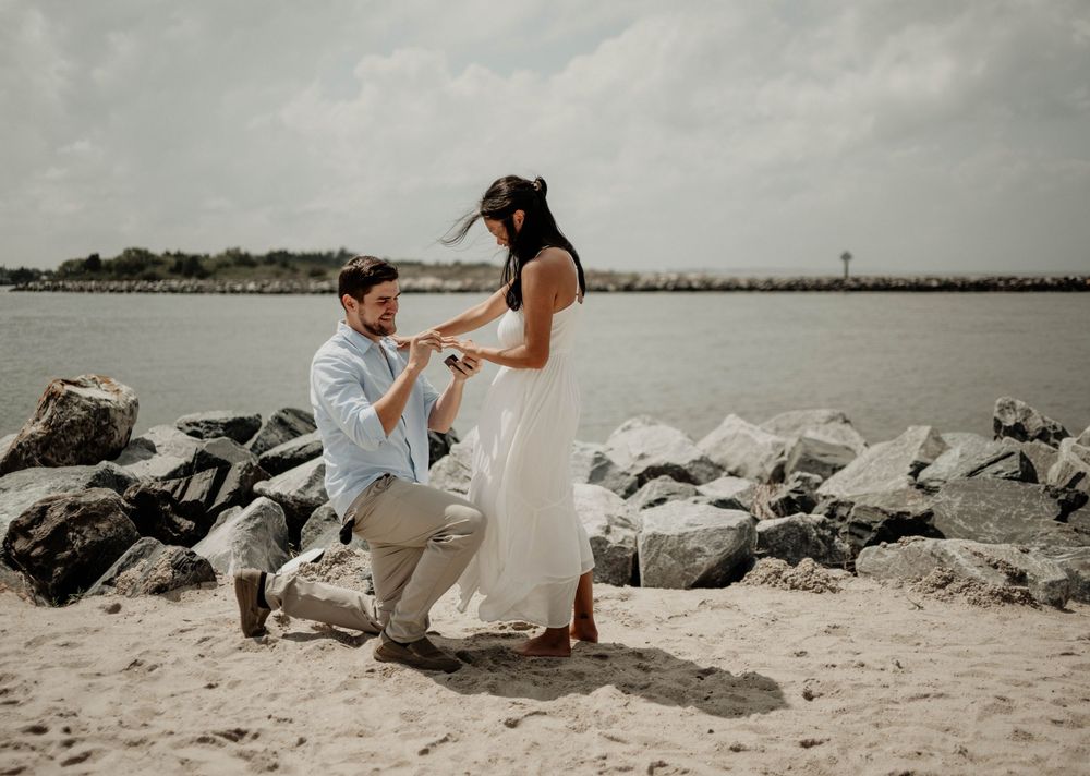 Man proposing to woman on sandy beach with rocky shoreline and ocean backdrop.
