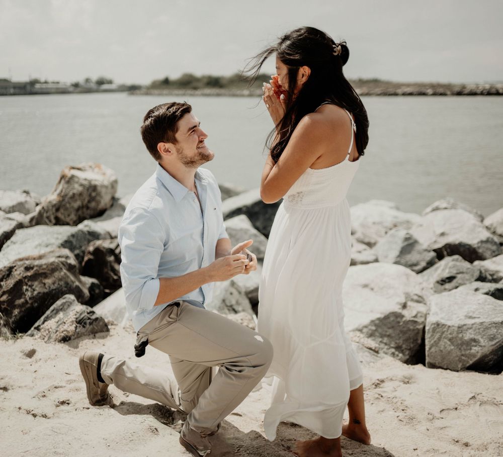 Man proposing on one knee to woman in white dress on sandy beach with rocky shoreline and water in background.