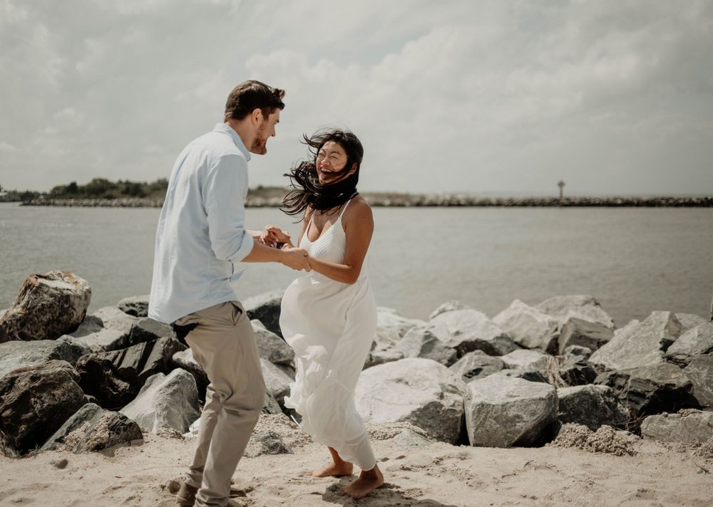 Couple dancing joyfully on a sandy beach near rocky jetty, woman in white dress, man in light blue shirt, cloudy sky.