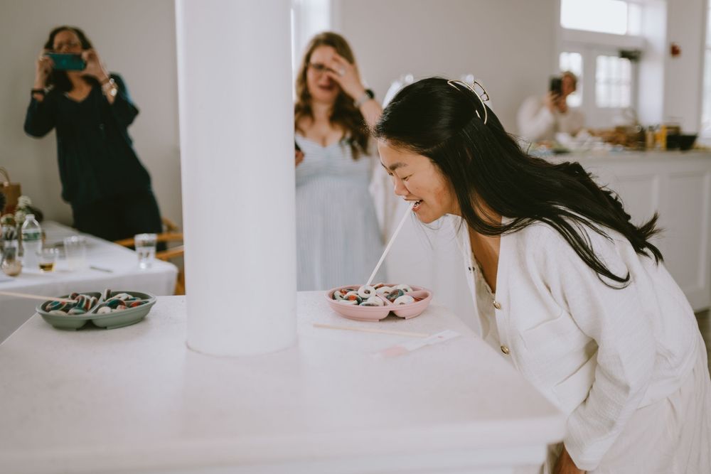 Bride in white robe leaning over cupcakes on table while photographer captures moment at wedding preparation.