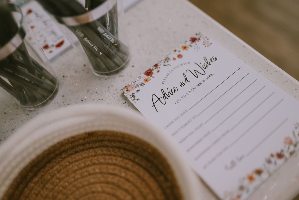 Floral-bordered advice and wishes card displayed on a table at a wedding reception with a woven basket nearby.