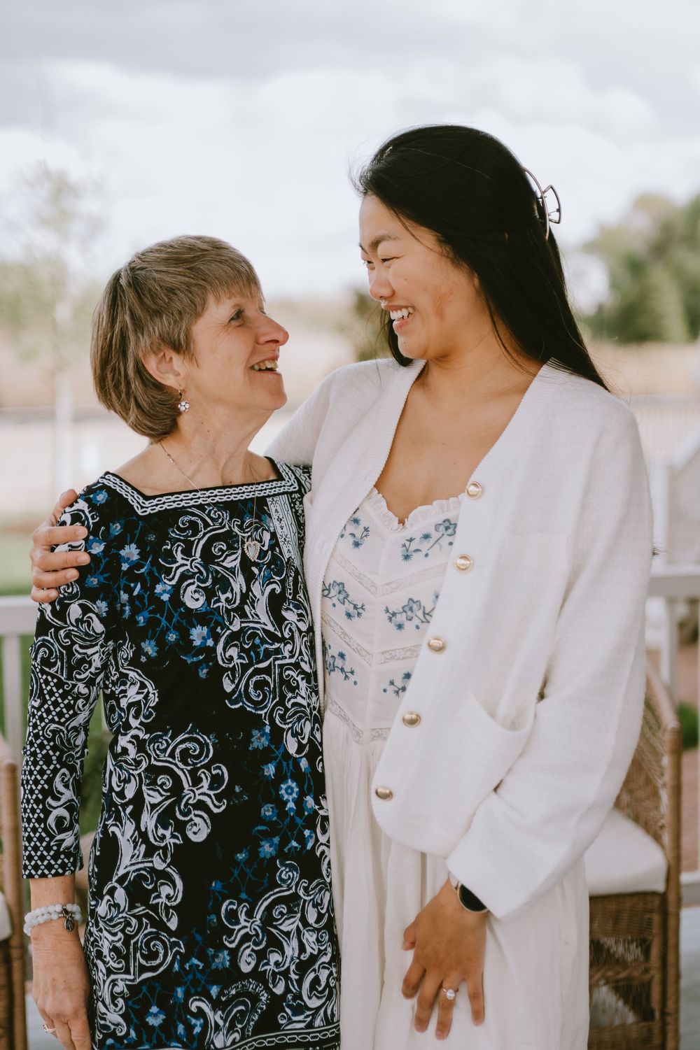 Two women smiling and embracing outdoors, one in a floral dress and one in a white outfit, sharing a joyful moment.