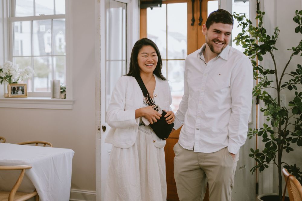 A smiling Asian woman and tall man stand together indoors in casual white attire near a window and green plant.
