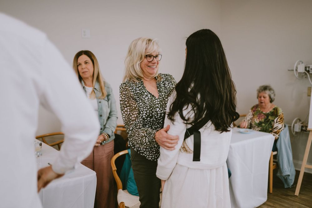 Women socializing and shaking hands at an indoor gathering, with guests seated at white-clothed tables in the background.