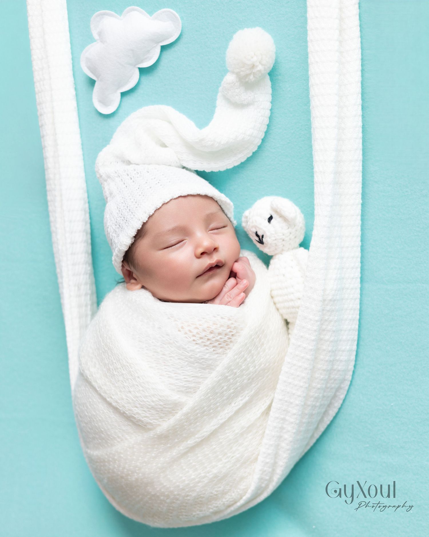 Newborn baby swaddled in white wrap wearing a knit hat, sleeping peacefully on a teal background with cloud decor.