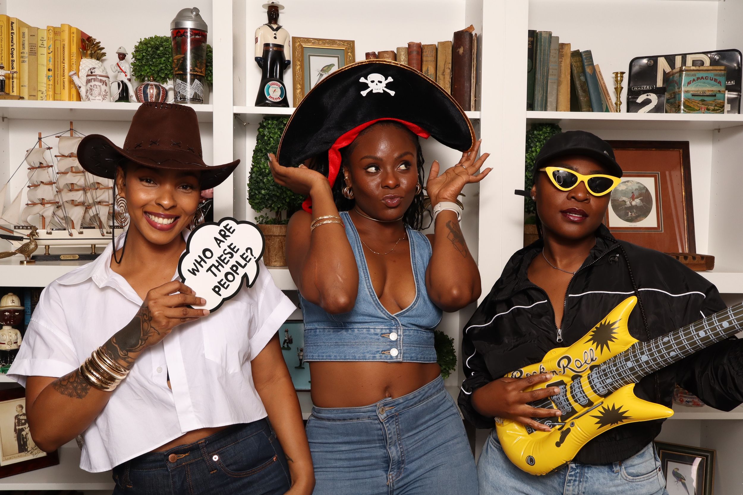 Three women pose playfully with pirate hat, cowboy hat, and toy guitar at a fun photo booth setup.