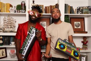 Two men pose with a keytar and boombox in front of white shelves filled with books and decor.