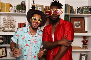Two stylish men pose wearing fun heart-shaped sunglasses in front of a white bookshelf display.