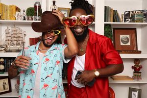 Two men laughing and posing with oversized red heart-shaped glasses and a cowboy hat at a fun indoor party.