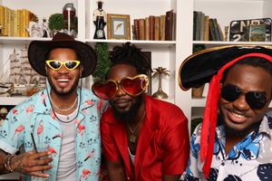 Three stylish men wearing colorful outfits and fun novelty sunglasses pose together smiling in a decorated room.