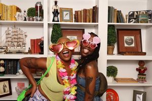 Two women wearing fun heart-shaped sunglasses and floral leis posing in front of a white bookshelf.