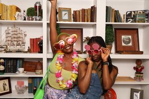 Two women wearing fun sunglasses and leis pose playfully in front of a bookshelf at a party.