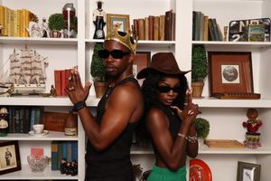 Two stylish friends posing back-to-back wearing a gold crown and brown fedora in front of a decorative bookshelf.