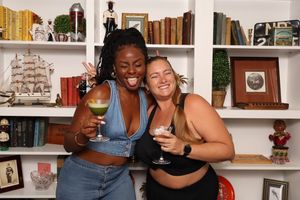 Two women smiling and holding cocktails in front of a white bookshelf filled with books and decor.