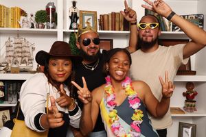 Four friends posing playfully with fun accessories including hats and a floral lei at a festive gathering.
