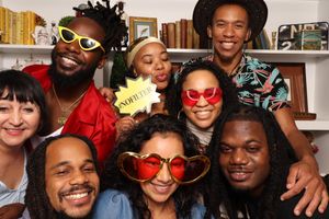 A diverse group of friends posing with fun props including oversized glasses and a novelty sign at a lively indoor party.