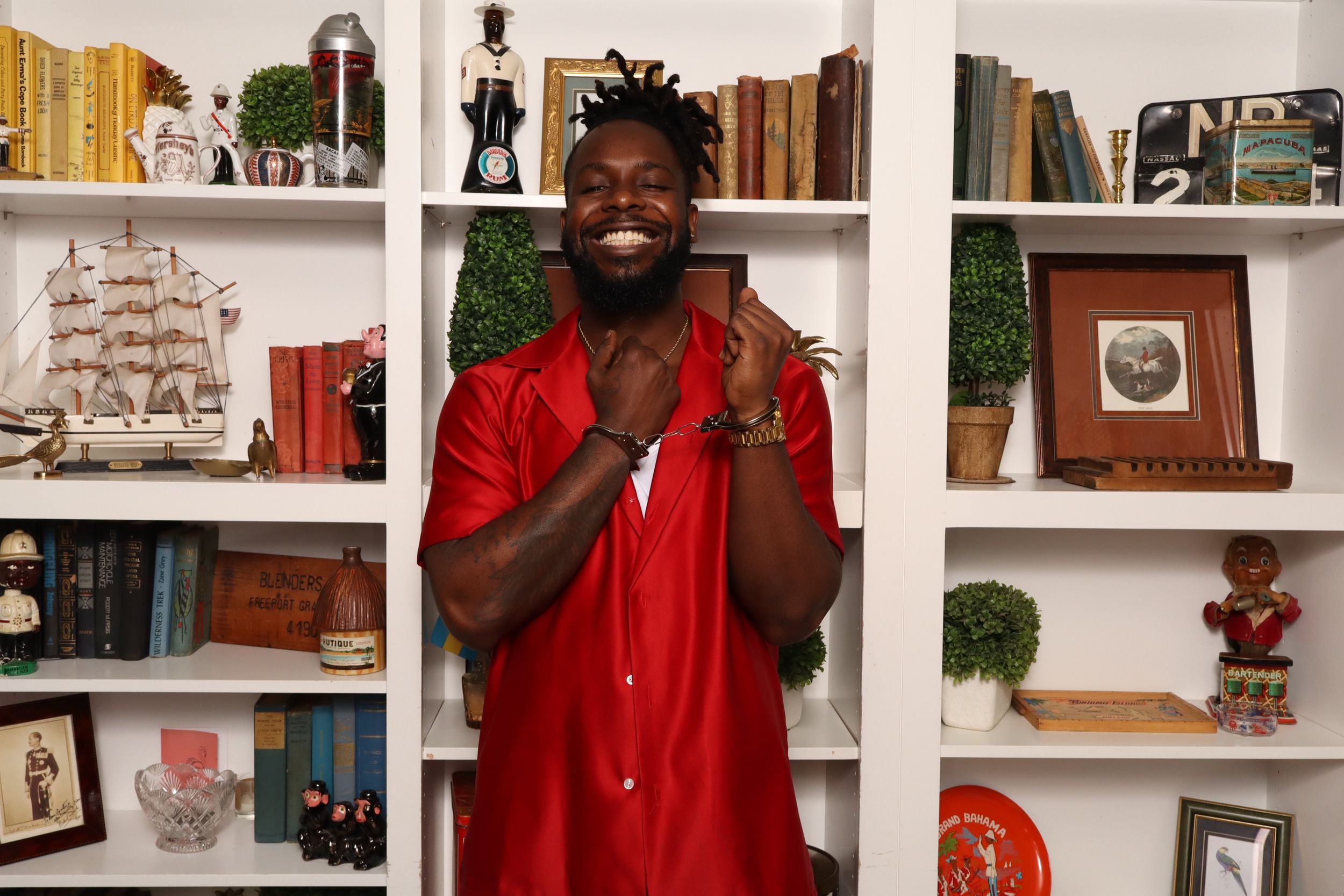 Man in red shirt smiling and clapping hands in front of white bookshelf with decorative items.