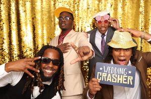 Four men pose playfully at a wedding photo booth with gold sequin backdrop, one holding a Wedding Crasher sign.