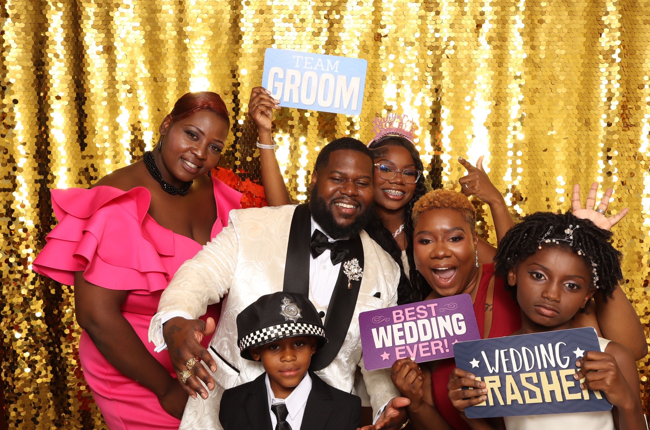 Group of joyful wedding guests posing with fun photo booth props including Groom and Best Wedding Ever signs against gold backdrop.