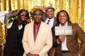 Four men posing at a photo booth with gold sequin backdrop, one holding an Open Bar sign at a festive event.