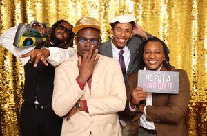 Four friends pose joyfully at a wedding photo booth with gold sequin backdrop, one holding a ring sign.