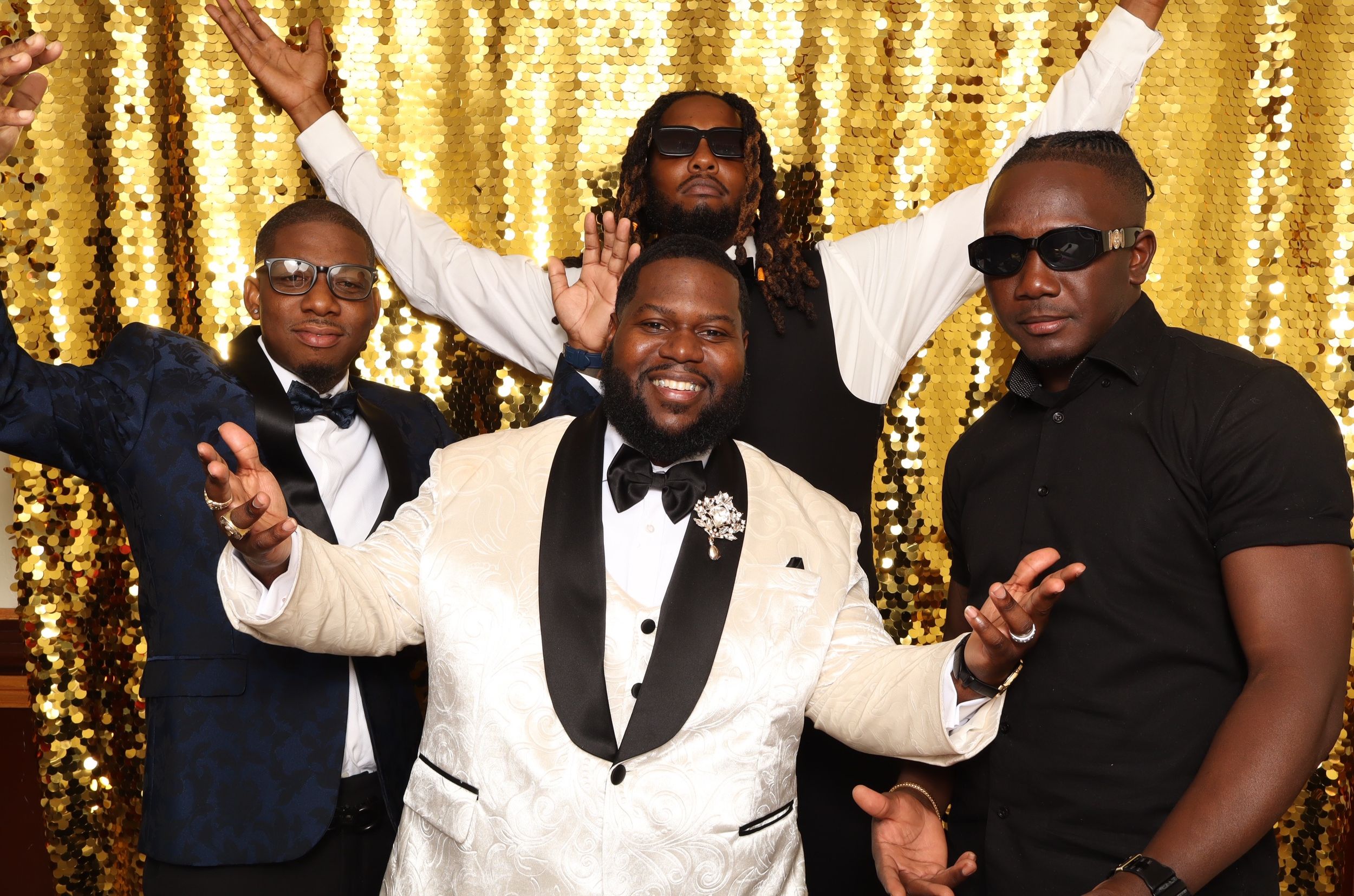 Four men posing and celebrating in front of a gold sequin backdrop at a formal event.