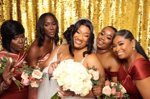 Bride in white gown holds bouquet surrounded by four bridesmaids in pink and red dresses against gold sequin backdrop.