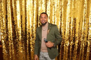 Smiling man in green blazer holding microphone in front of gold sequin backdrop.